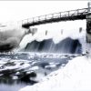 A snow-covered dam with cascading water under a metal footbridge in a winter landscape