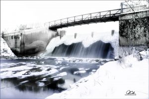 A snow-covered dam with cascading water under a metal footbridge in a winter landscape