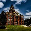 Historic red-brick courthouse with clock tower under a partly cloudy sky, surrounded by green lawn and trees