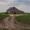 An old, dilapidated barn tilts noticeably on an overgrown path surrounded by lush green fields under a cloudy sky.