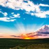 A vibrant sunset with sun rays over rolling green fields and a bright blue sky with scattered clouds.