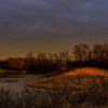 A tranquil landscape with bare trees surrounding a serene pond under a dramatic, cloudy sky, and the last rays of sunlight casting a warm glow on the grass.