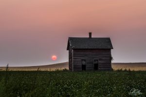 Old wooden house in a field with a red sun setting behind it, under a pink sky.
