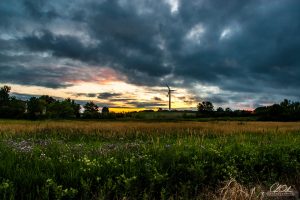 A wind turbine under a dramatic sky with a field in the foreground at sunset.