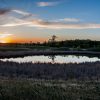 A serene sunset over a small pond surrounded by tall grasses and distant trees.