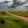 Dirt road winding through lush green rolling hills under a dramatic stormy sky with swirling dark clouds.