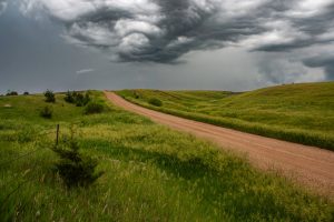 Dirt road winding through lush green rolling hills under a dramatic stormy sky with swirling dark clouds.