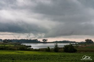 A stormy landscape with dark clouds looming over a serene lake and lush green fields.