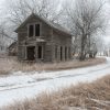 An abandoned wooden house stands amid snow-covered ground and barren trees with a winding path leading away.