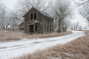 An abandoned wooden house stands amid snow-covered ground and barren trees with a winding path leading away.