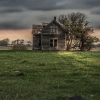 An abandoned wooden house stands solitary in a grassy field under a clouded sky with distant trees.