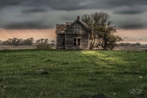 An abandoned wooden house stands solitary in a grassy field under a clouded sky with distant trees.