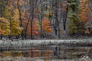 A tranquil pond reflects the vibrant autumn foliage of orange, red, and yellow, dusted with a light snowfall in a serene forest setting.