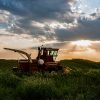 A lone red tractor rests in a green field under a dramatic sky with sun rays breaking through clouds at sunset.