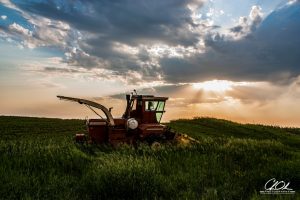 A lone red tractor rests in a green field under a dramatic sky with sun rays breaking through clouds at sunset.