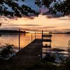 Wooden dock leading to a calm lake during a vibrant sunset with trees framing the scene.