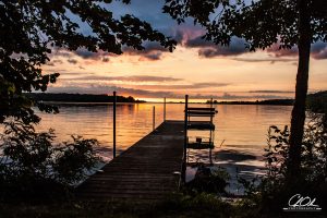 Wooden dock leading to a calm lake during a vibrant sunset with trees framing the scene.