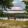 A wooden bench facing a serene lake, framed by trees and reeds, with sunlight breaking through the clouds in the distance.