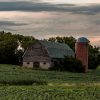 An old barn with attached silo surrounded by lush green fields and trees under a cloudy sky.