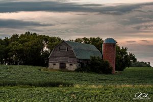 An old barn with attached silo surrounded by lush green fields and trees under a cloudy sky.