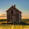 An old wooden house stands isolated in a sunlit field with a sky transitioning from blue to golden hues.