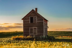 An old wooden house stands isolated in a sunlit field with a sky transitioning from blue to golden hues.