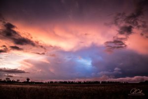 A vast sky at sunset painted with vibrant hues of pink, orange, and blue above a darkened silhouette of a horizon line with distant trees.