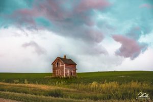 An old wooden farmhouse stands alone in a vast green field beneath a picturesque sky with colorful, swirling clouds.