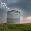 Lightning strikes near a solitary grain silo in a vast green field under a stormy sky.