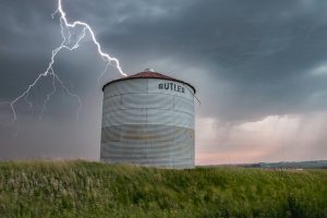 Lightning strikes near a solitary grain silo in a vast green field under a stormy sky.