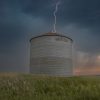 Grain bin with lightning strike in open field under dark stormy sky.