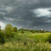 A landscape with dark storm clouds looming over lush green fields and scattered trees