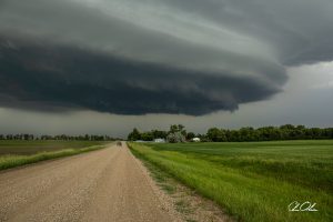 Gravel road leading towards a distant farm under a dark, looming storm cloud.
