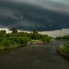 Dark storm clouds looming over a river with lush green trees on either side.