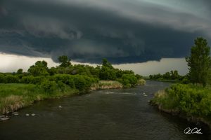 Dark storm clouds looming over a river with lush green trees on either side.