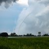 A car driving on a highway with a tornado funnel in the distance against a cloudy sky.