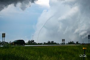 A car driving on a highway with a tornado funnel in the distance against a cloudy sky.