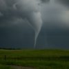A dark, swirling tornado touching down on a green prairie under stormy skies.