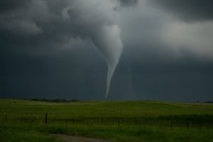 A dark, swirling tornado touching down on a green prairie under stormy skies.
