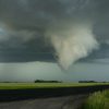 A formidable tornado funnel cloud descending from dark storm clouds over a flat rural landscape, with a vehicle on the right and a field of green grass stretching to the horizon.