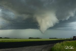 A formidable tornado funnel cloud descending from dark storm clouds over a flat rural landscape, with a vehicle on the right and a field of green grass stretching to the horizon.