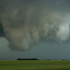 A large, ominous cloud formation resembling a funnel cloud hangs over a flat, green field under a dark sky.