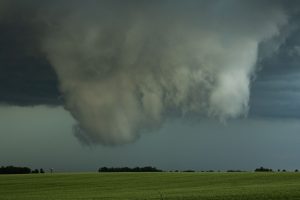 A large, ominous cloud formation resembling a funnel cloud hangs over a flat, green field under a dark sky.