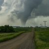 A narrow dirt road leads past green fields toward a distant tornado under swirling storm clouds.