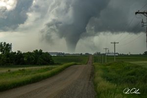 A narrow dirt road leads past green fields toward a distant tornado under swirling storm clouds.