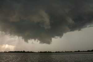 Large dark storm clouds above a body of water with a distant horizon of trees.