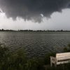 A dark storm cloud over a calm lake with a white bench by the grassy shore.