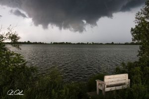 A dark storm cloud over a calm lake with a white bench by the grassy shore.