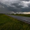 A tornado forms under dark storm clouds over a rural road and grass fields.