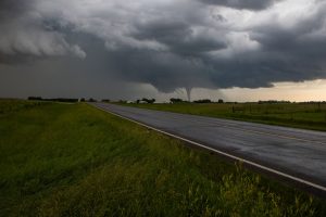 A tornado forms under dark storm clouds over a rural road and grass fields.